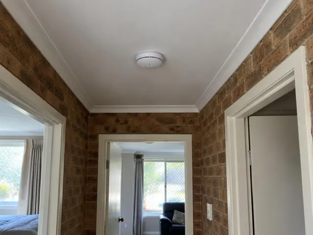 Ceiling-mounted smoke alarm in a hallway with brick walls and white trim in Arcadia Vale